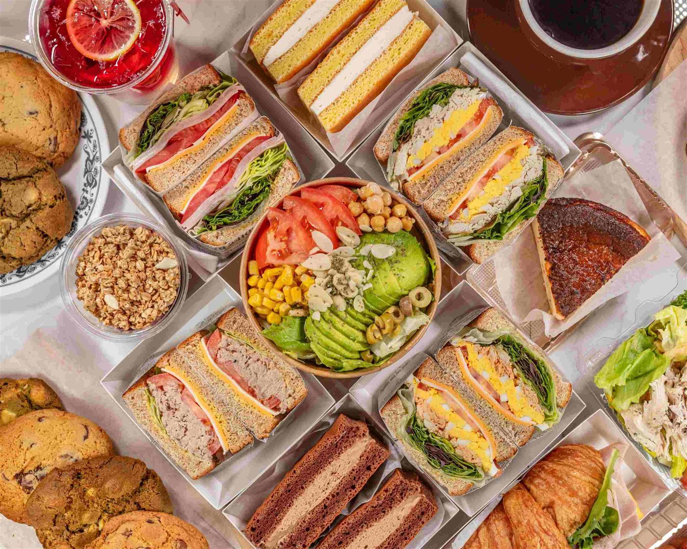 Overhead spread of Zagmachi sandwiches, an avocado salad bowl, cookies, Basque cheesecake and drinks